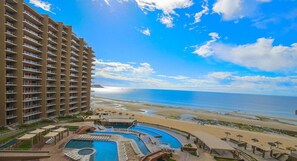 Un hotel de gran altura con varios balcones que dan a una playa y un área de piscina con múltiples piscinas y tumbonas.