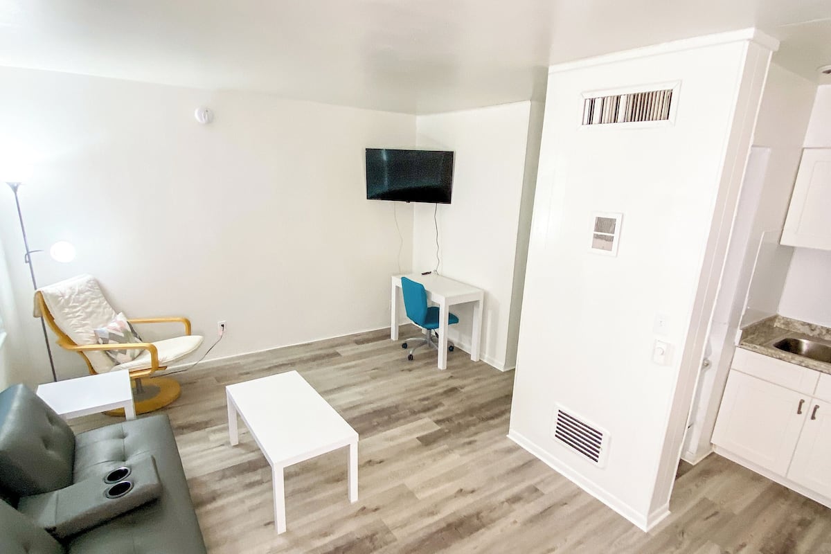 A modern living room with a grey sofa, a white coffee table, a blue office chair, a flat-screen TV mounted on the wall, and a kitchenette area with white cabinets and a sink.
