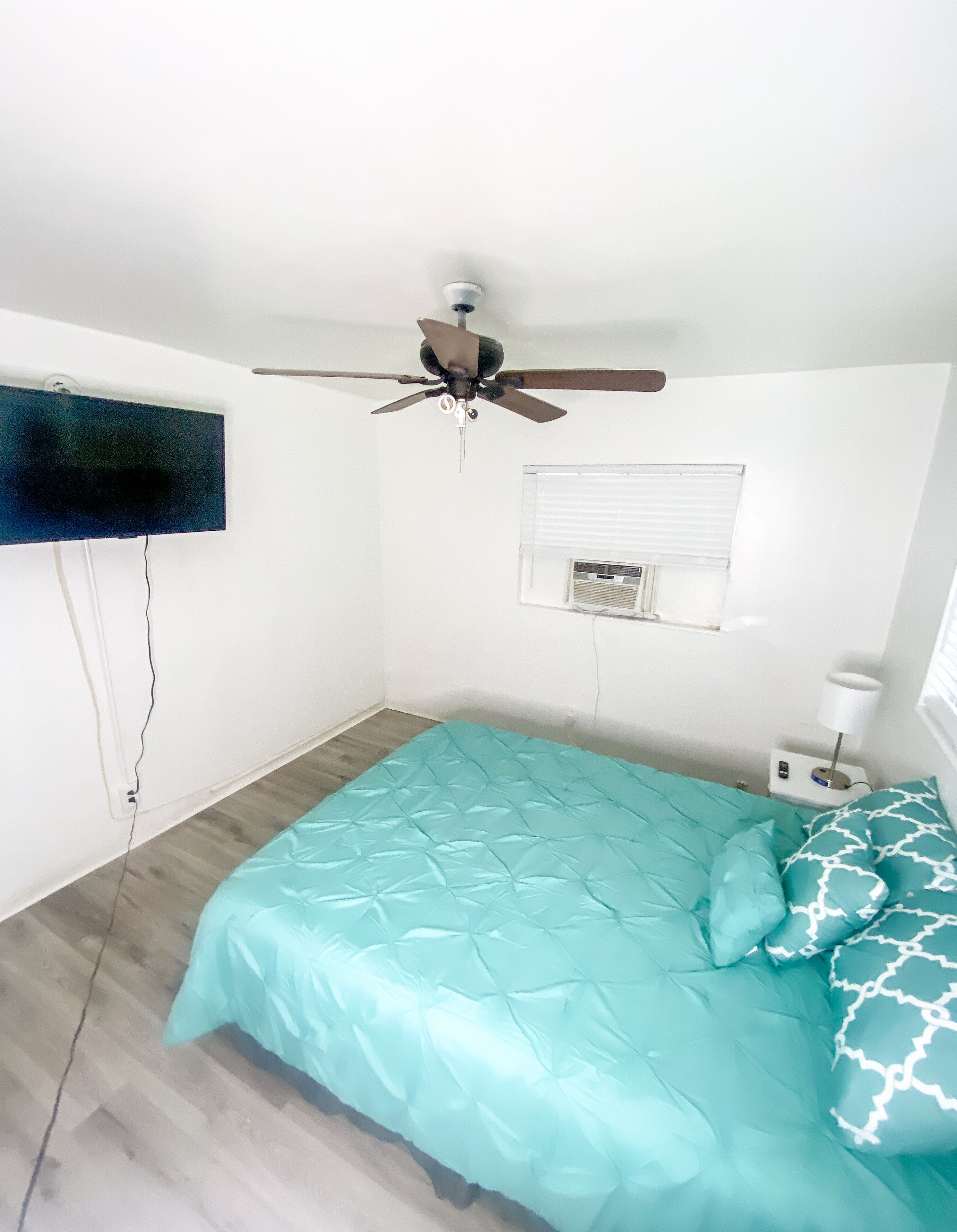 A bedroom with a bed, ceiling fan, TV, air conditioning unit, and bedside lamp.