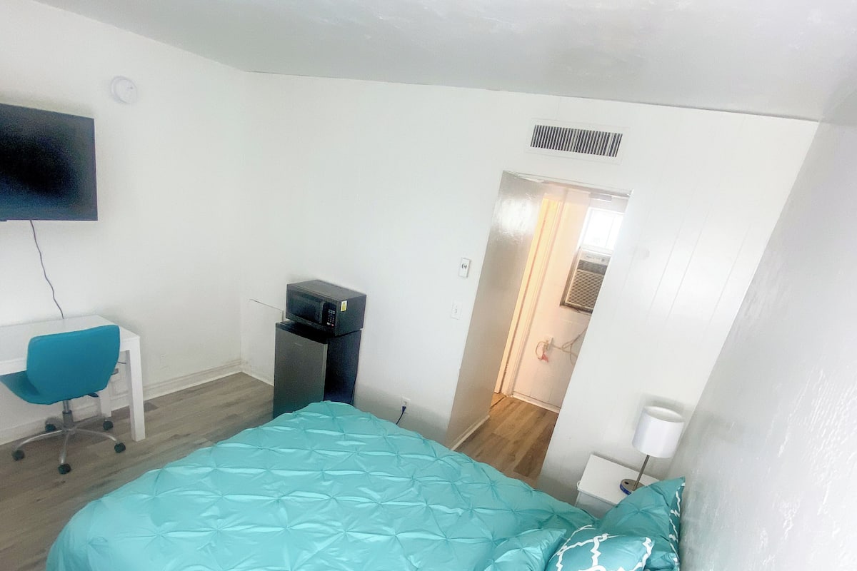 A bedroom with a bed, a desk, a chair, a TV, and a door leading to another room.