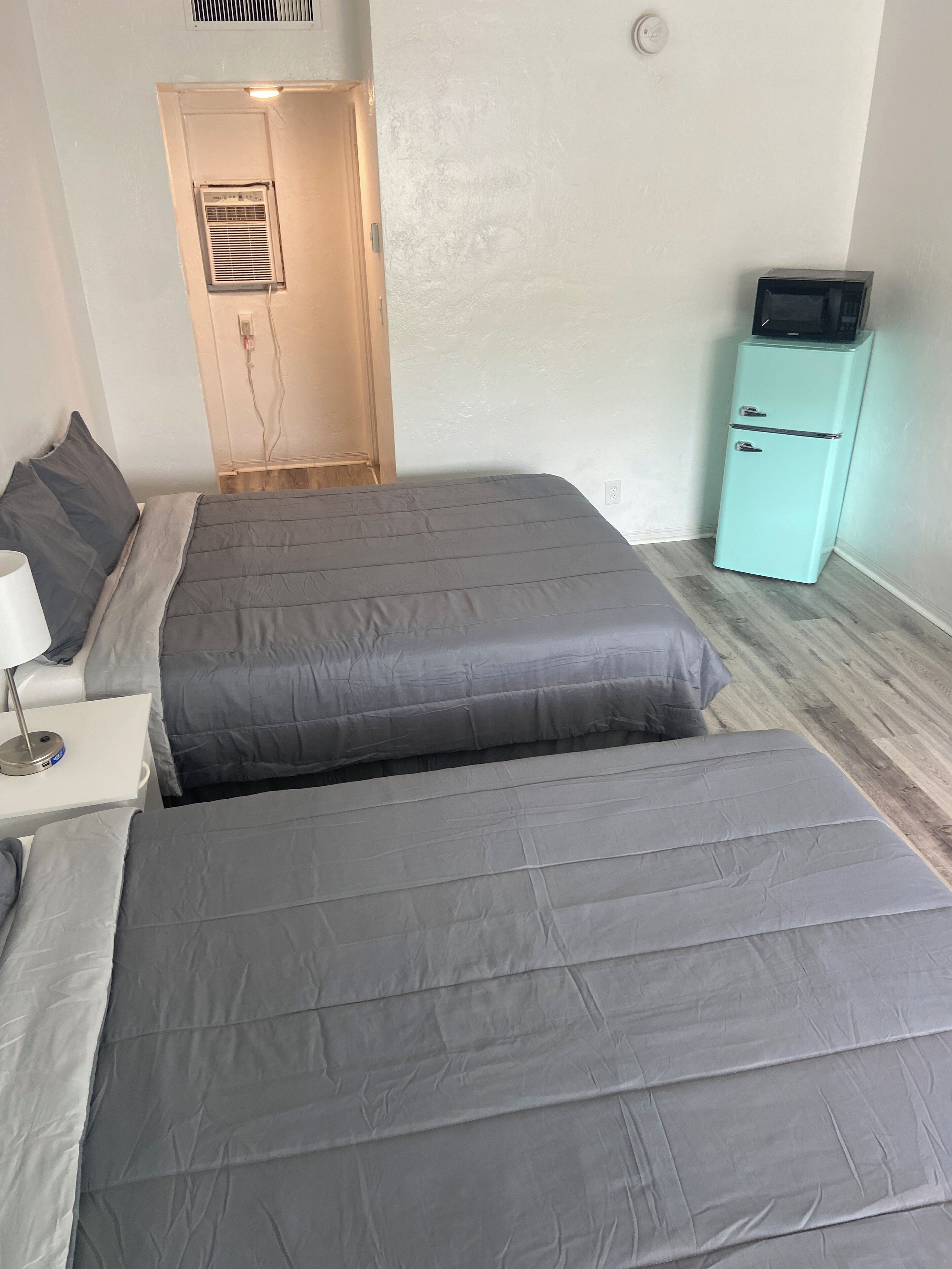 Un dormitorio con dos camas, una mesita con lámpara, un refrigerador color turquesa y una puerta que da a otra habitación.