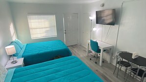 A room with two beds, a desk, a chair, a TV, and a window with blinds. - Uptown Suites (St. Petersburg)