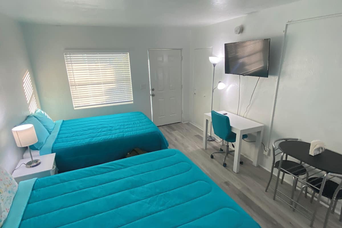 A room with two beds, a desk, a chair, a TV, and a window with blinds.