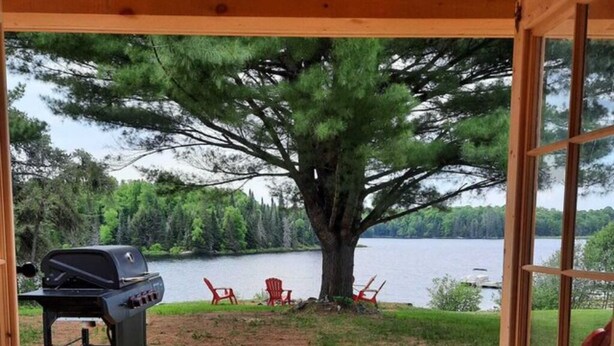 A wooden deck with red Adirondack chairs, a grill, and a view of a lake and forest. - Site #45 | Blue Lake Bunkie (Whitestone)