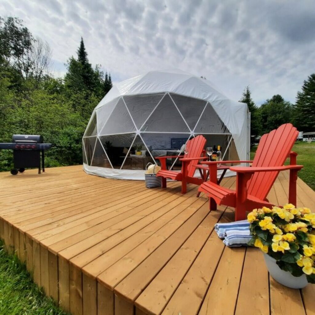 A geodesic dome tent with a wooden deck, red Adirondack chairs, and a grill.
