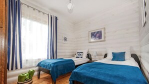 A room with two beds, a wooden floor, a window with curtains, a framed picture on the wall, and a small green basket on the floor. - Nice apartment in Jaroslawiec with WiFi (Jaroslawiec)