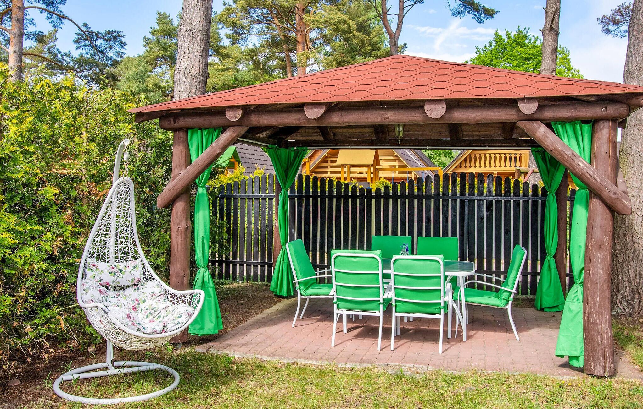 A wooden gazebo with a red roof, surrounded by a black picket fence. Inside, there are green chairs and a white table.