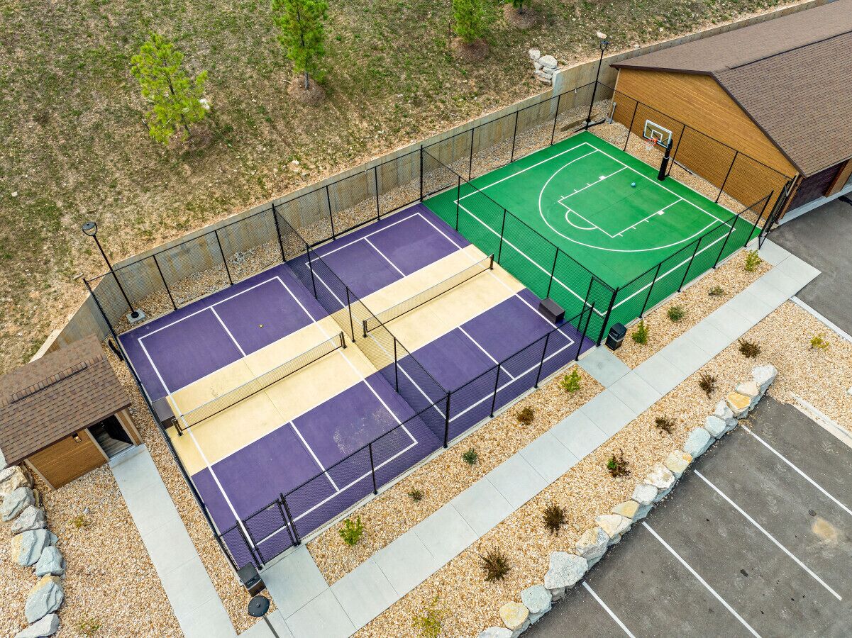 Un terrain de basket en plein air, doté d’une surface de jeu verte et violette, entouré d’une clôture et d’un petit bâtiment.