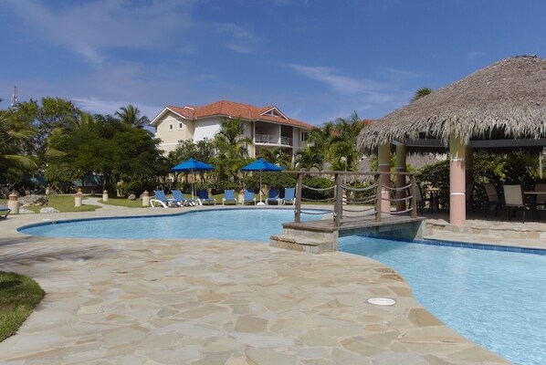 A pool area with lounge chairs, umbrellas, and a thatched-roof structure.