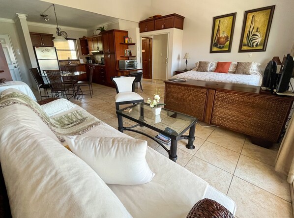 A living room with a sofa, coffee table, and a dining area with chairs. There is a kitchen visible in the background.