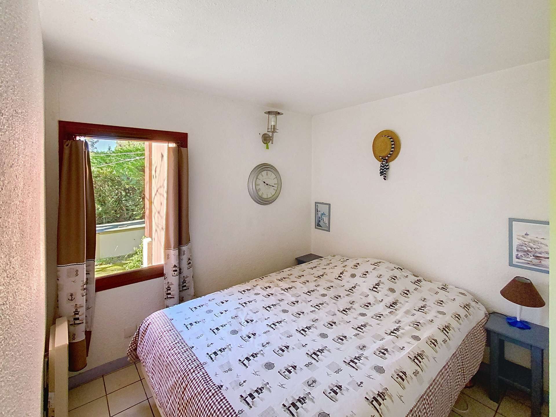 Een slaapkamer met een bed, een raam, een wandklok en een klein tafeltje met een lamp.