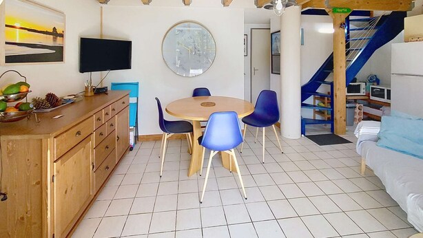 A room with a wooden ceiling, a round table with chairs, a television, a clock, and a staircase with a blue railing.