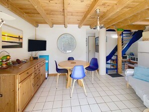 A room with a wooden ceiling, a round table with chairs, a television, a clock, and a staircase with a blue railing. - 196 – Holiday Home for 6 People (HOURTIN)
