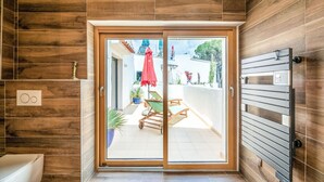 Ein modernes Badezimmer mit Holzverkleidung, eine Glastür zu einer Terrasse im Freien mit Tisch, Stühlen und einem roten Sonnenschirm.