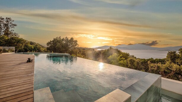 Ein Infinity-Pool mit Holzterrasse, umgeben von üppiger Vegetation und einem malerischen Blick auf einen Sonnenuntergang über Wasser und Berge.