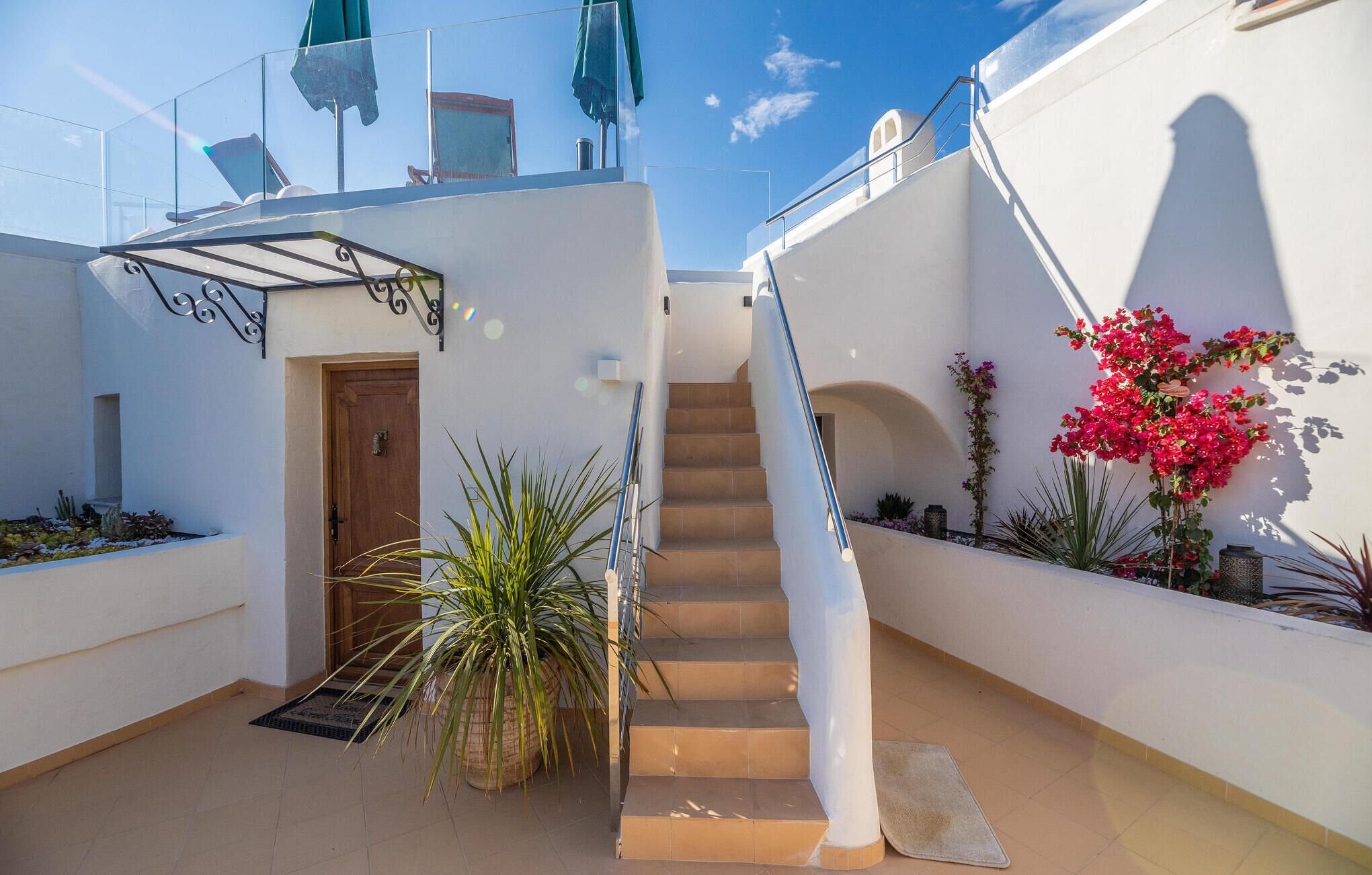 Un escalier extérieur moderne menant à un balcon du deuxième étage, doté d’une rambarde en verre, et entouré de plantes en pot, avec un ciel dégagé en toile de fond.