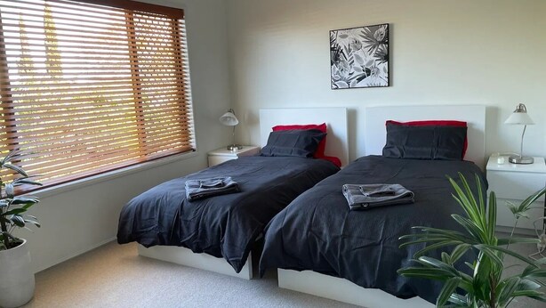 A bedroom with two beds, a window with blinds, a wall-mounted artwork, bedside tables with lamps, and potted plants. - Our Townhouse in Toowoomba (Newtown)