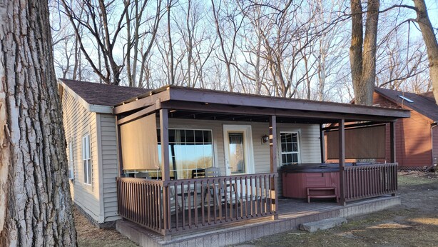 A small, single-story cabin with a porch, a hot tub, and a covered outdoor seating area. - Riverfront Cottage with Hot Tub, Kayaks, Canoes, & Fishing Boats! (Waterville)