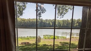 A view through a window with a metal grid, overlooking a lake with a grassy area and trees in the background. - Riverfront Cottage with Hot Tub, Kayaks, Canoes, & Fishing Boats! (Waterville)