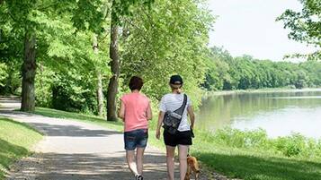 Two people walking on a paved path with a dog, surrounded by lush green trees and a body of water.