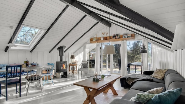 Un loft espacioso con techo alto, vigas a la vista, un tragaluz grande, estufa de leña, zona de comedor y un rincón de estar acogedor con sofá y cojines.