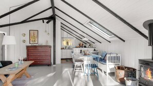 Un loft espacioso con techo alto, vigas a la vista, comedor con mesa y sillas de madera, cocina con armarios blancos, sofá azul y estufa de leña.