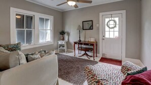 A living room with a ceiling fan, a fireplace, a door with a wreath, and a window with curtains. - The Carriage House Downtown Lafayette, LA (Lafayette)