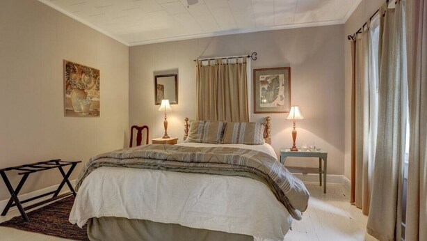 A bedroom with a large bed, two bedside tables with lamps, a wooden chair, and artwork on the walls. - The Carriage House Downtown Lafayette, LA (Lafayette)