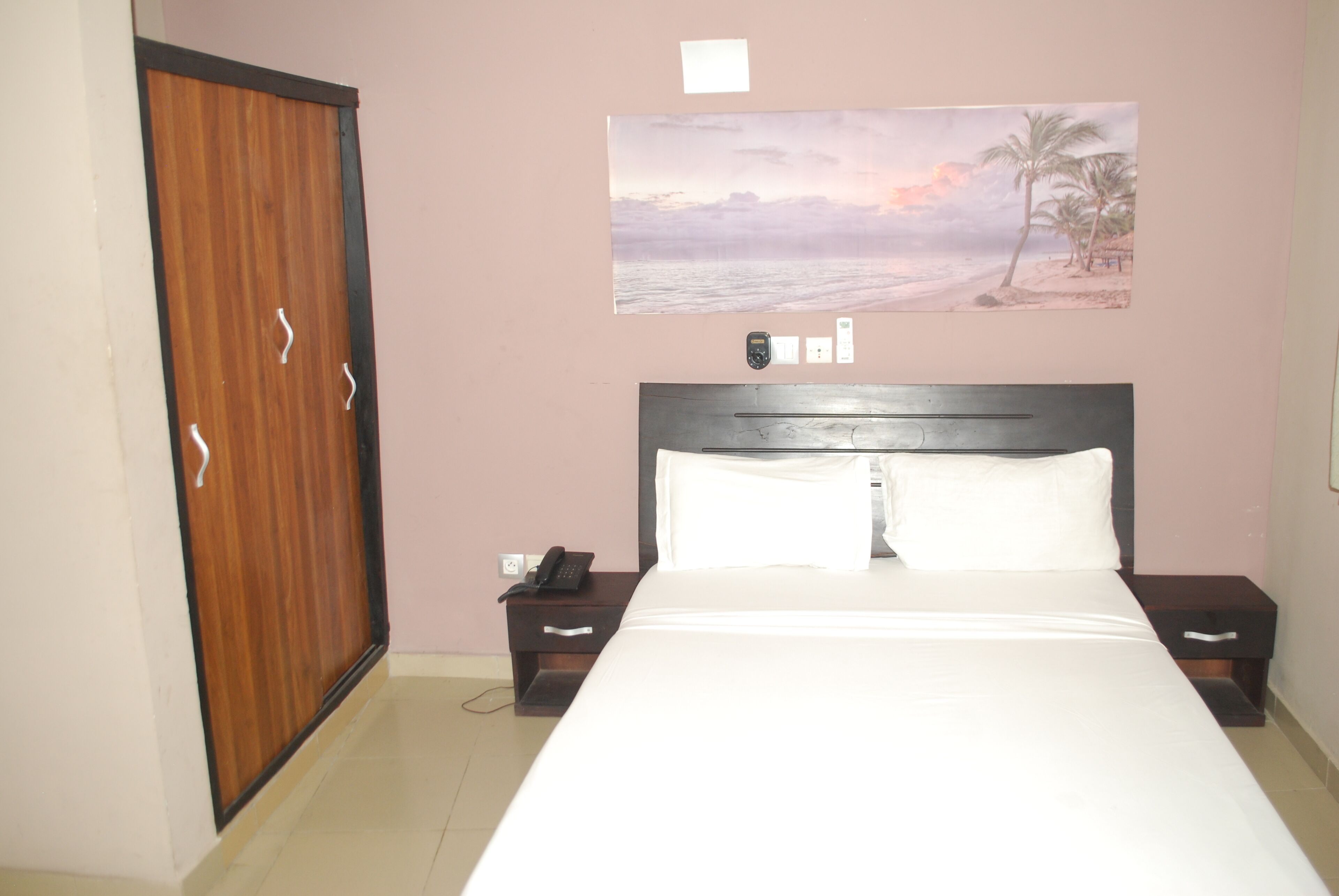 A bedroom with a bed, a nightstand, a phone, a wall-mounted light switch, and a door.