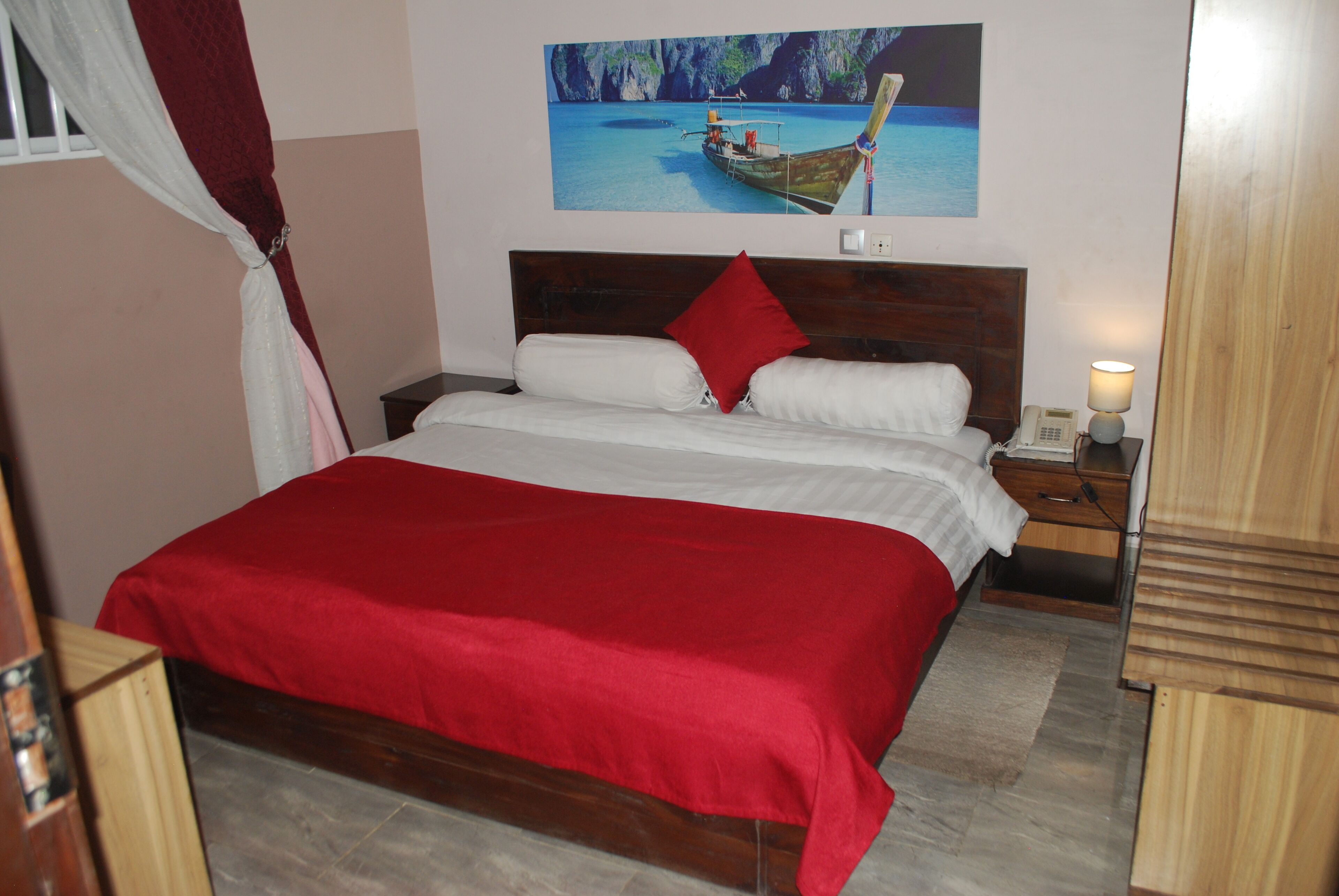 A bedroom with a wooden bed, red bedding, a nightstand with a lamp, and a painting of a boat on the wall.