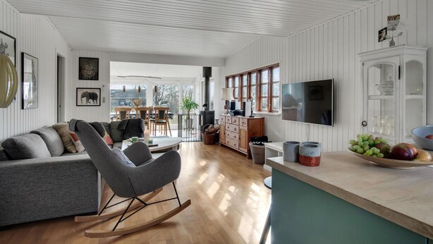 A modern living room with a grey sofa, a rocking chair, and a wooden floor. There is a kitchen area with a TV, cabinets, and a fruit bowl. - Beautiful home in Grevinge with kitchen (Grevinge)