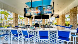 A spacious bar area with high-top tables and blue cushioned chairs, a large flat-screen TV mounted above the bar, and a view of palm trees and buildings in the background.