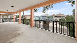 Ein überdachter Balkon mit Betonboden, schwarzem Metallgeländer und Blick auf Wohnhäuser und Palmen.