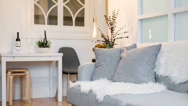 Un salon moderne avec un canapé gris, un plaid blanc et moelleux, et un coin repas comprenant une table et des chaises blanches.