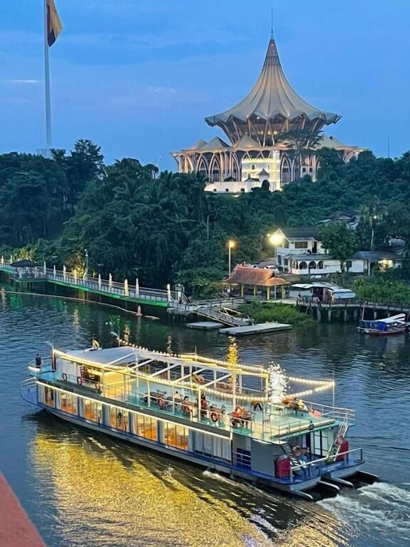 A river with a boat and a large building with a unique roof structure. - Natol Homestay-Riverbank Suites High Rise City Centre (Kuching)