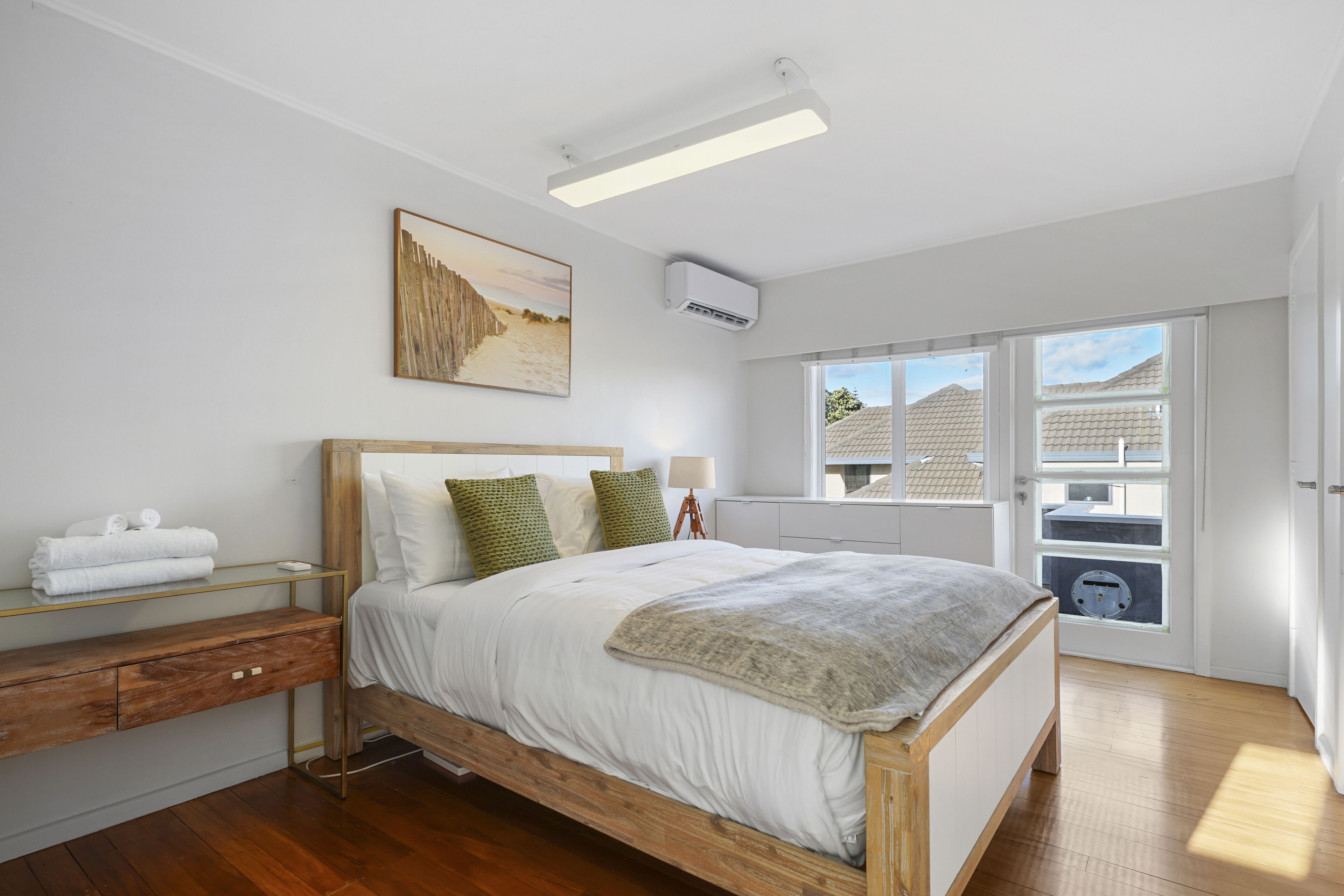 A bedroom with a wooden bed, white bedding, a bedside table with towels, a wall-mounted air conditioner, and a window with a view of buildings.