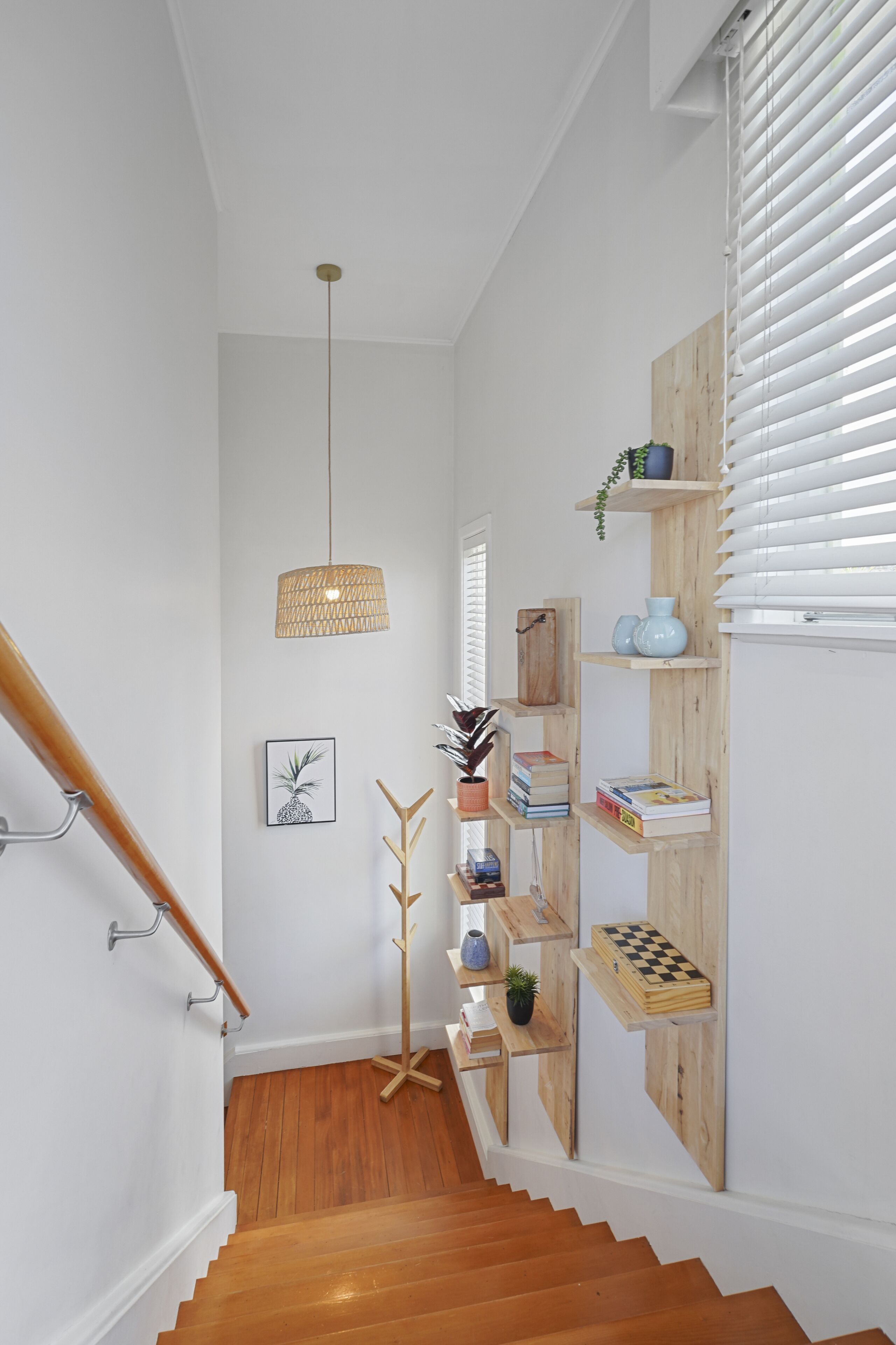 Eine Treppe mit Holzstufen und einer Handläufe, die zu einer Zwischenebene führt, auf der eine Pendelleuchte aus Rattan, Holzregale mit Büchern und Dekorationsgegenständen sowie ein gerahmtes Bild an der Wand zu sehen sind.