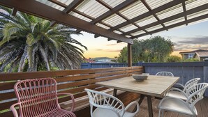A covered outdoor patio with a wooden table and white chairs, surrounded by a wooden fence and palm trees. - Amazing Home By Beach - Aircon-ed  (Auckland)