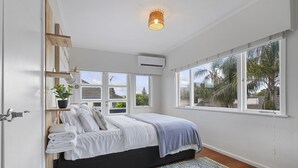 A bedroom with a bed, a bedside table with a plant, a window with a view of trees, and an air conditioning unit. - Amazing Home By Beach - Aircon-ed  (Auckland)
