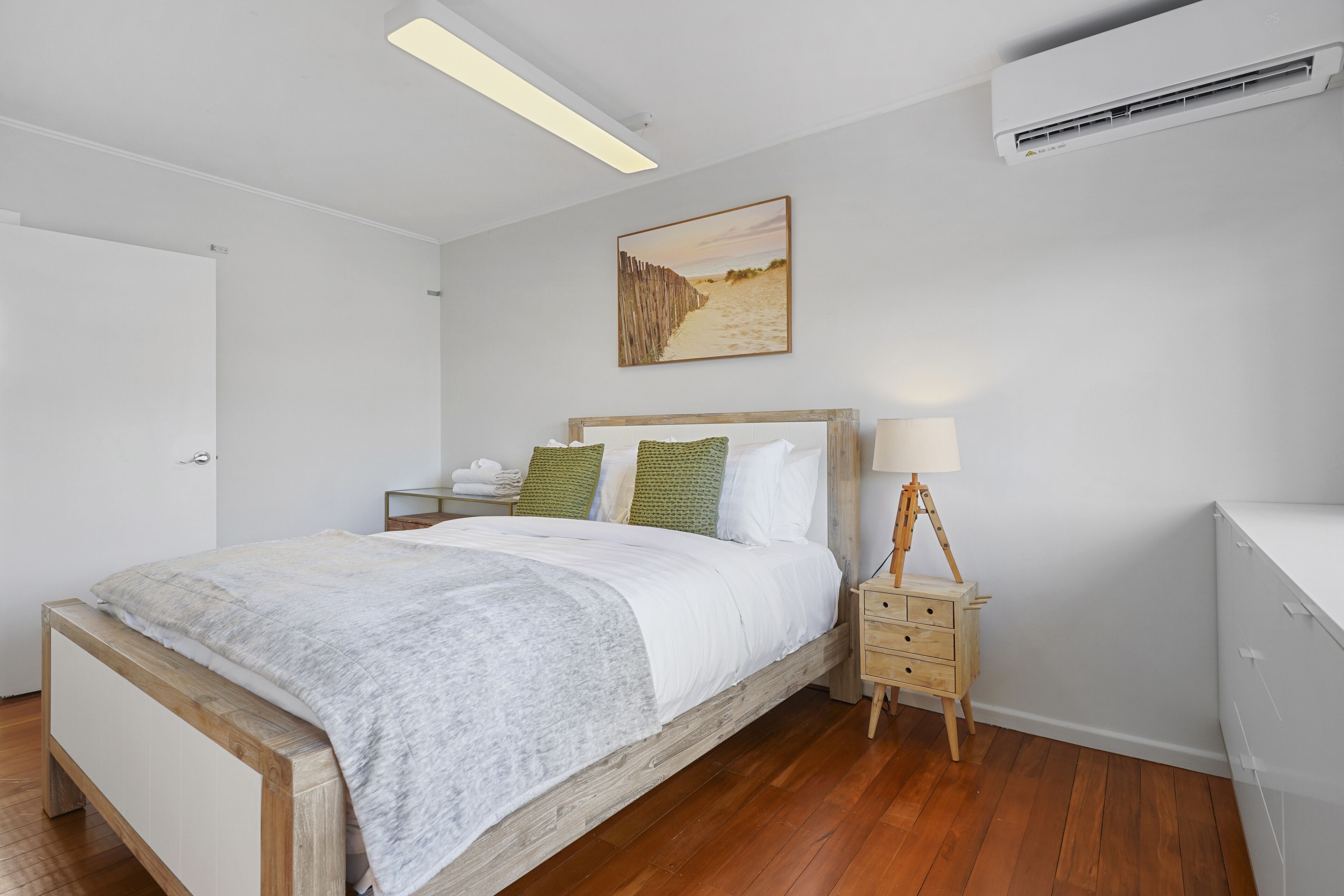 A bedroom with a wooden bed, white bedding, a nightstand with a lamp, and an air conditioning unit on the wall.