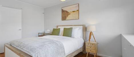 A bedroom with a wooden bed, white bedding, a nightstand with a lamp, and an air conditioning unit on the wall.
