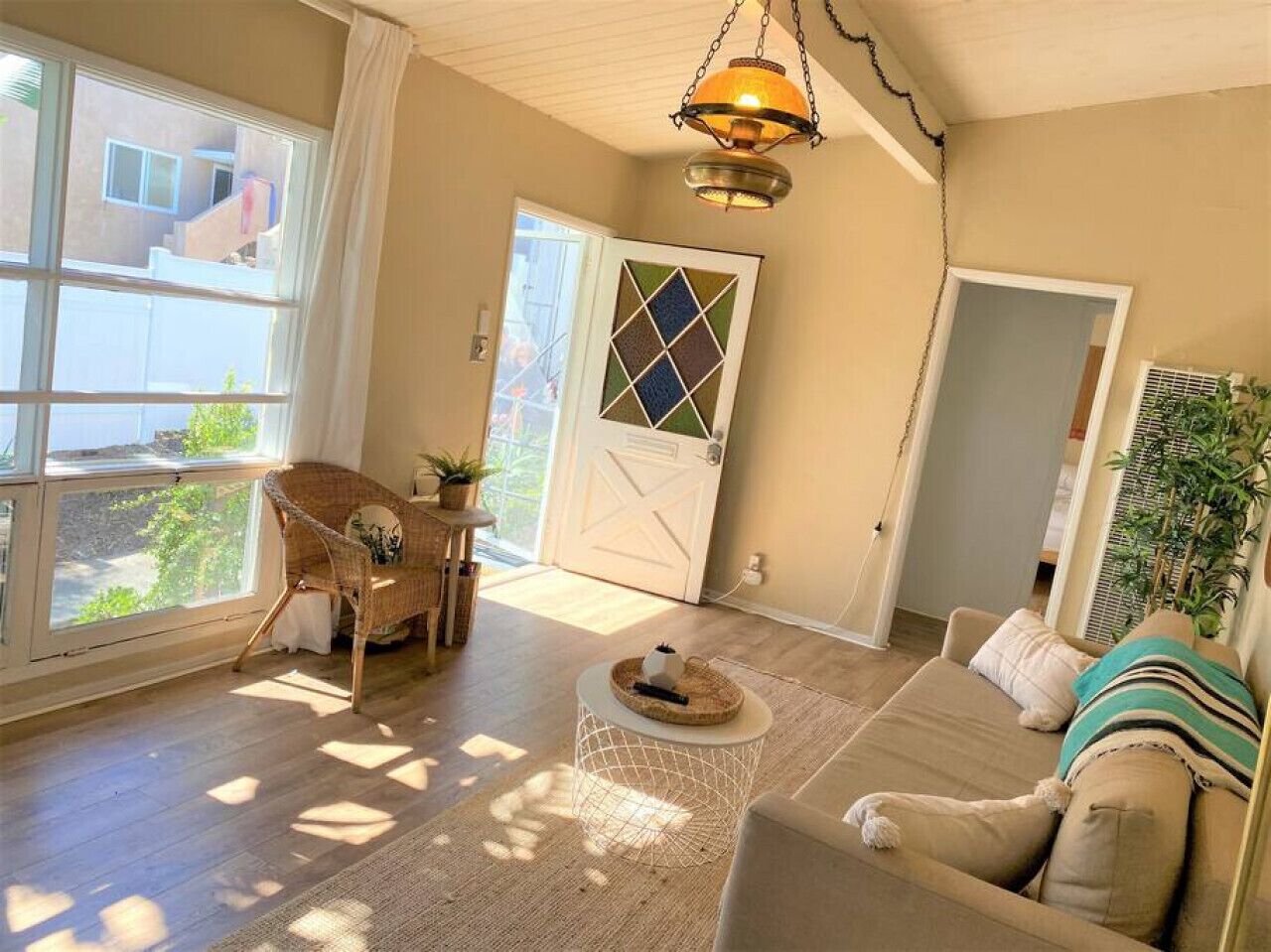 A spacious living room with a large window, a wicker chair, a sofa, and a hanging light fixture.