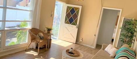 A spacious living room with a large window, a wicker chair, a sofa, and a hanging light fixture.