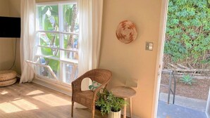 Quarto com cadeira de vime, uma pequena mesa redonda, uma planta em vaso e vista para o verde através de uma porta de vidro.