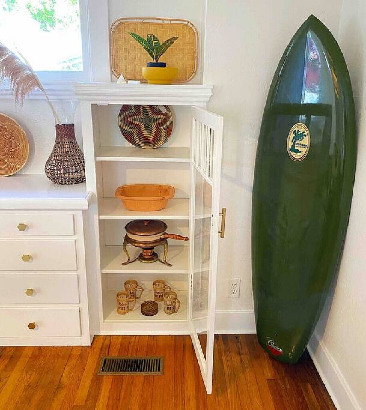 A room with a white cabinet containing kitchenware, a surfboard, and decorative items.