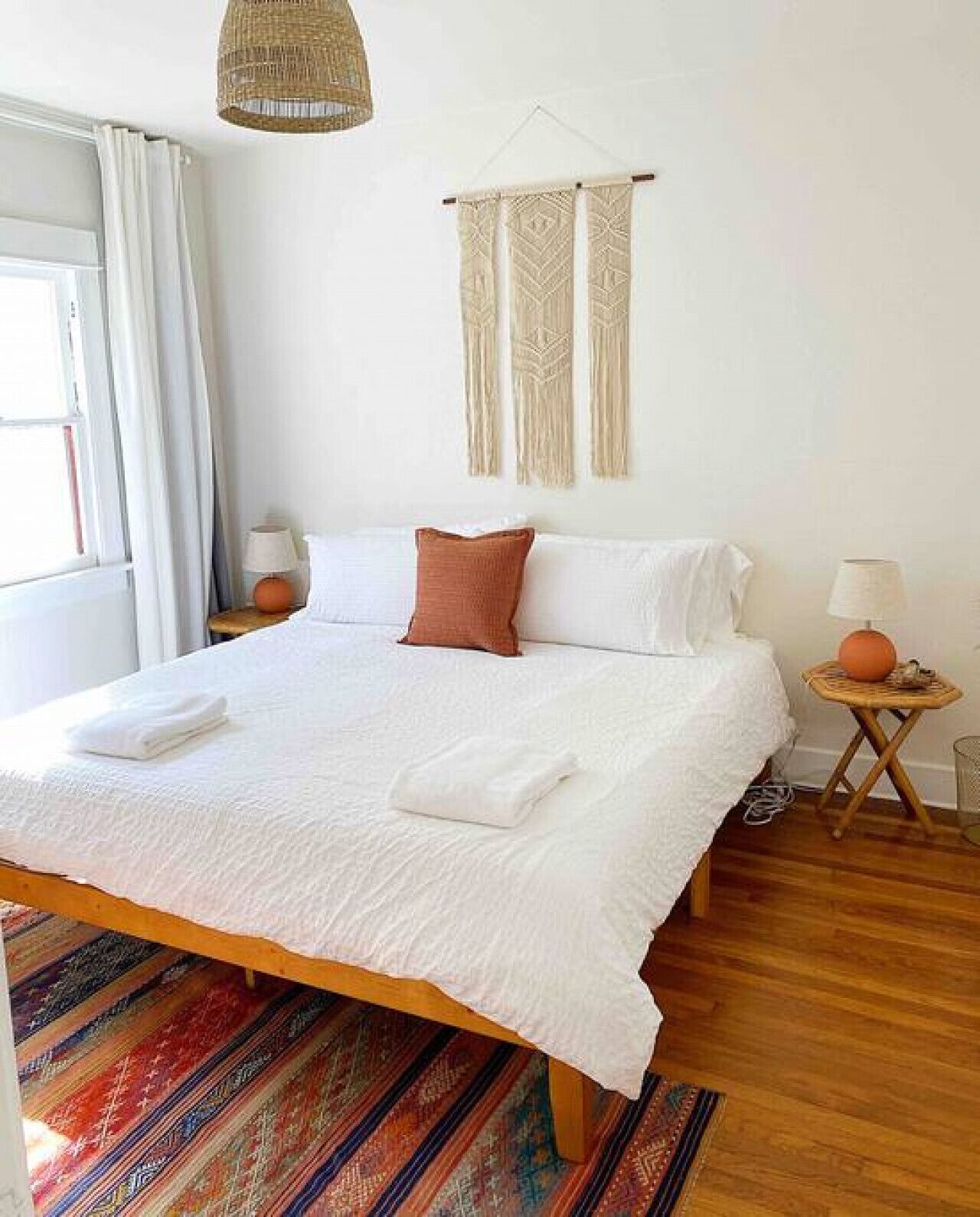 A neatly made bed with white linens and a single brown pillow, a woven wall hanging, and a small wooden side table with a lamp.