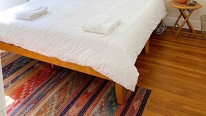 A neatly made bed with white linens and a single brown pillow, a woven wall hanging, and a small wooden side table with a lamp.