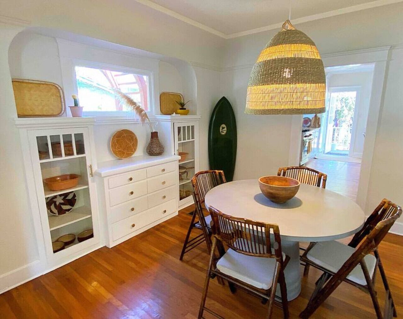 A dining area with a round table, wooden chairs, a hanging light fixture, and a surfboard on the wall.