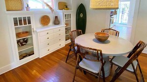 A dining area with a round table, wooden chairs, a hanging light fixture, and a surfboard on the wall.