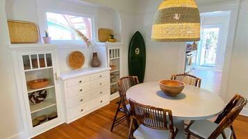 A dining area with a round table, wooden chairs, a hanging light fixture, and a surfboard on the wall.